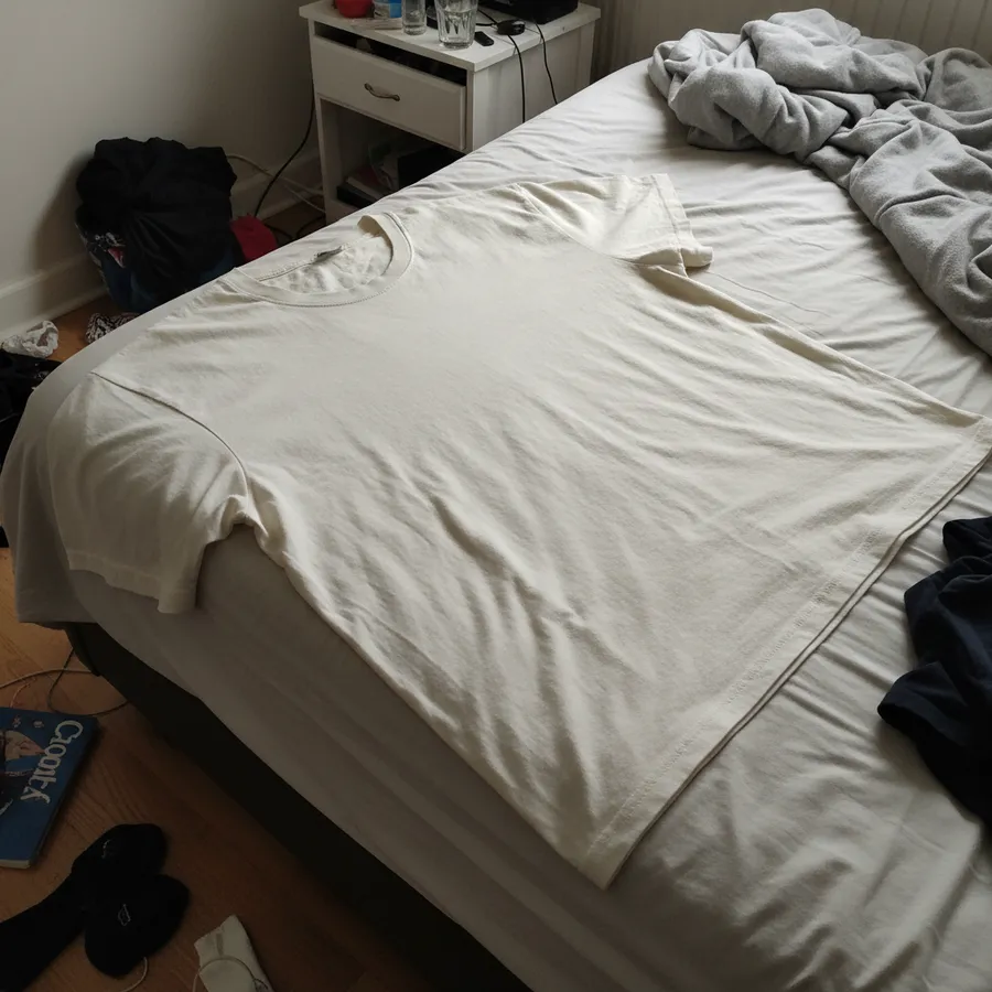 Camiseta off-white dobrada em cima de uma cama desarrumada, fotografada com o celular sob luz amarelada de quarto